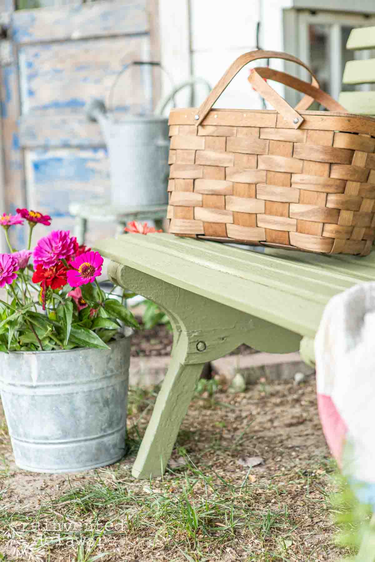A green painted wooden bench with a woven picnic basket on it stands next to a galvanized metal bucket filled with pink flowers. The background includes additional rustic outdoor elements, such as a weathered wooden door and another metal bucket.