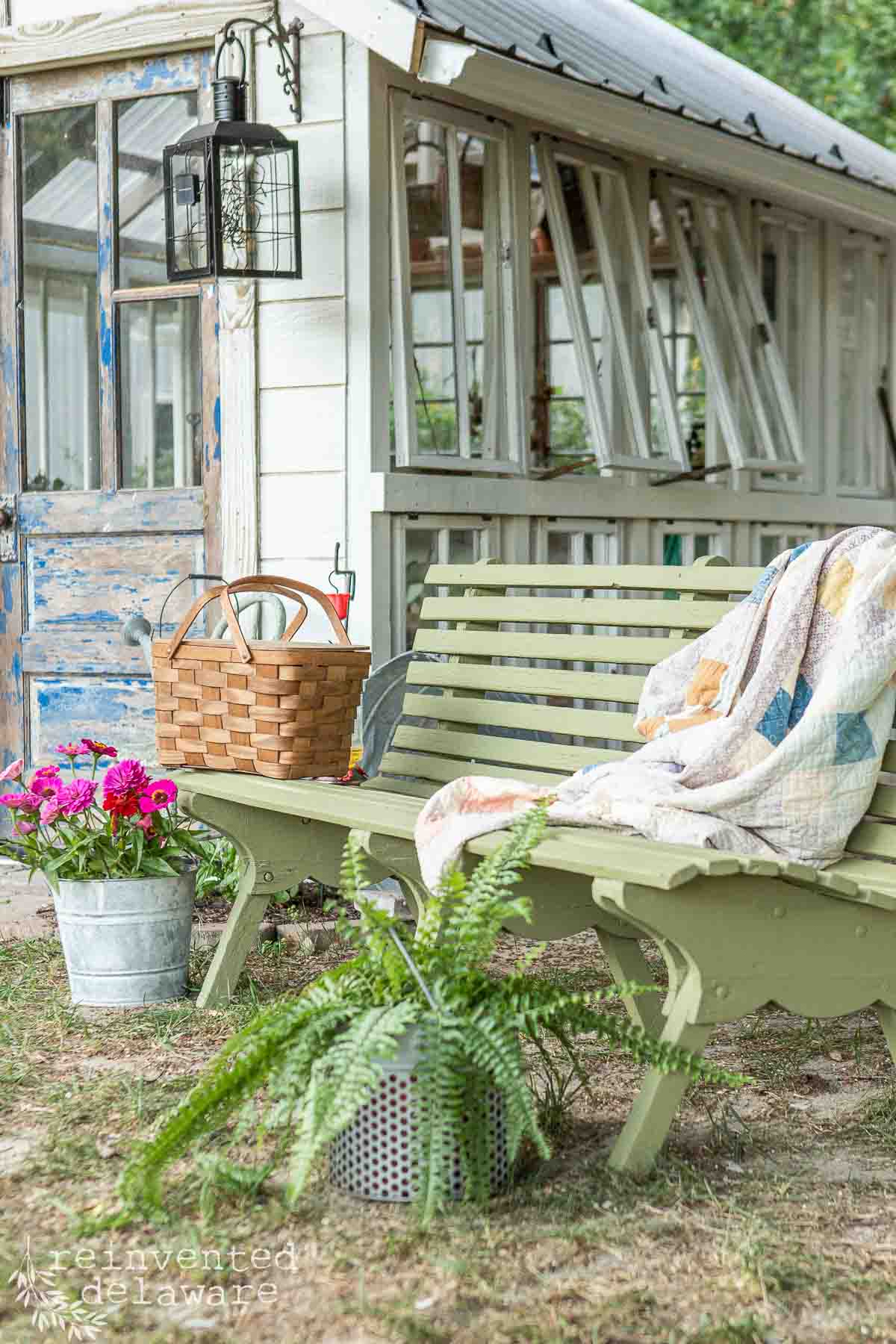 A cozy garden scene features a green wooden bench draped with a quilt and accompanied by a bucket of pink flowers and a potted fern. Nearby, a rusted metal lantern hangs, and a wicker basket sits on the bench. A quaint she shed greenhouse is in the background.