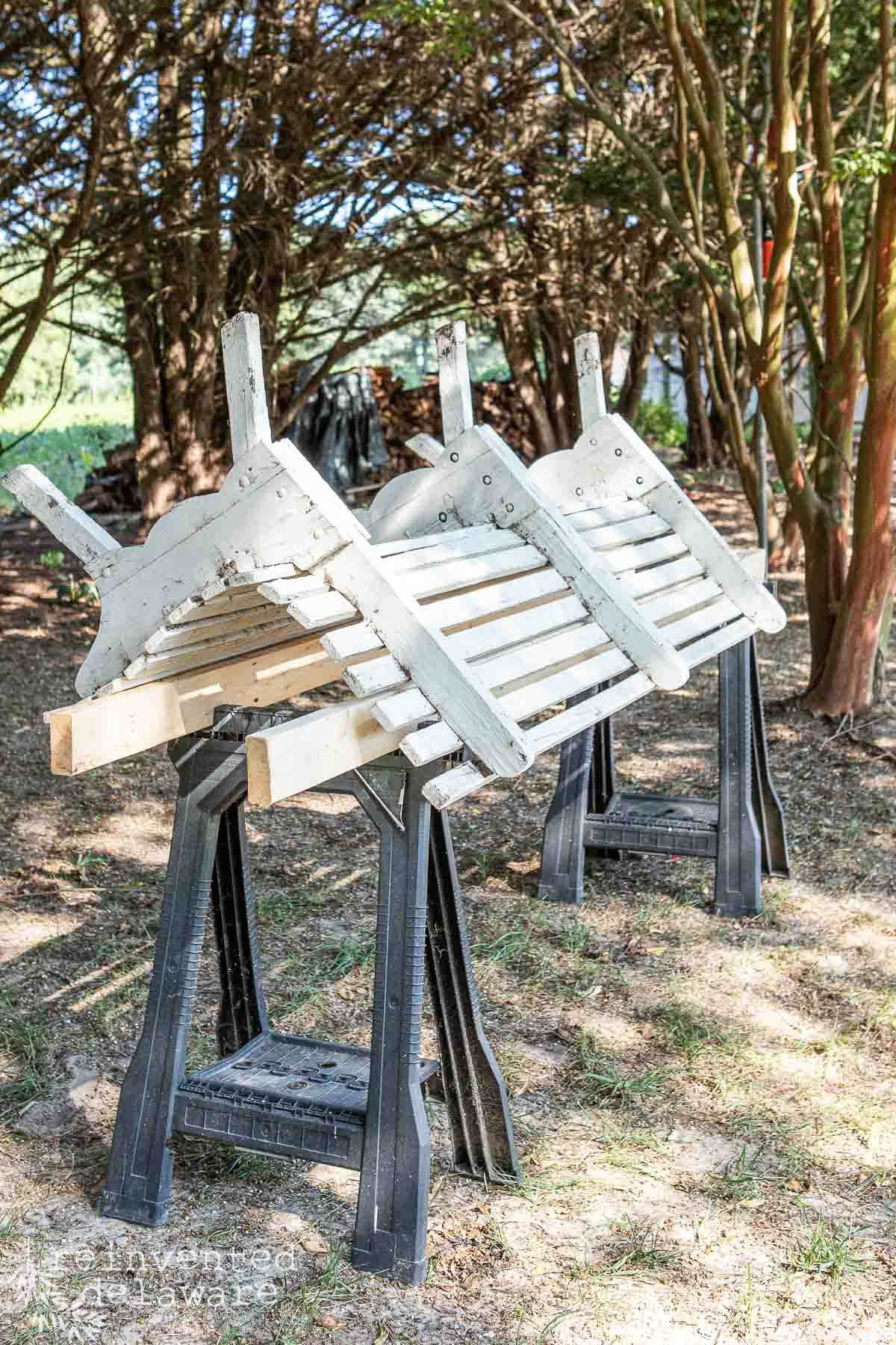 Three white wooden bench outdoors on black sawhorses. The surrounding area features trees and green foliage, casting dappled shadows on the ground. The scene appears to be in a garden or park setting.