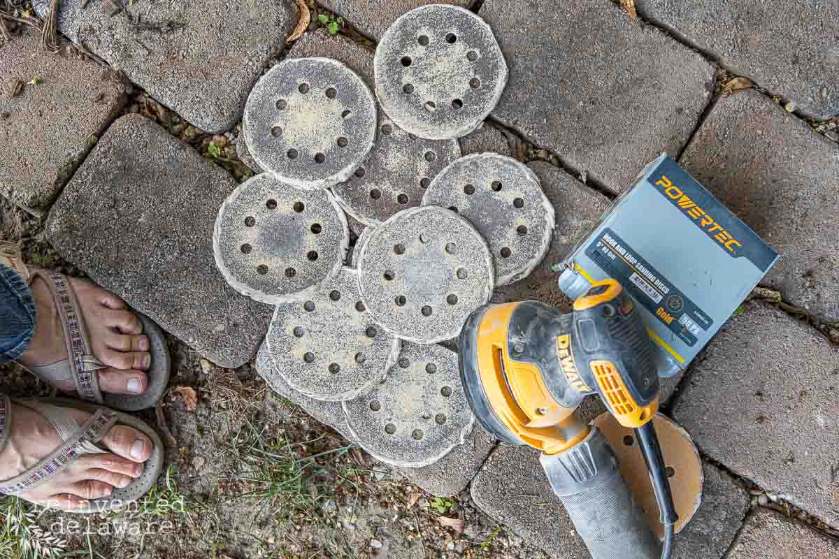 A person stands next to a DeWalt orbital sander, several used circular sandpaper discs with multiple holes, and a Powertec Sanding Disc box on a stone-paved surface. The scene indicates recent sanding work, likely on wooden outdoor furniture.
