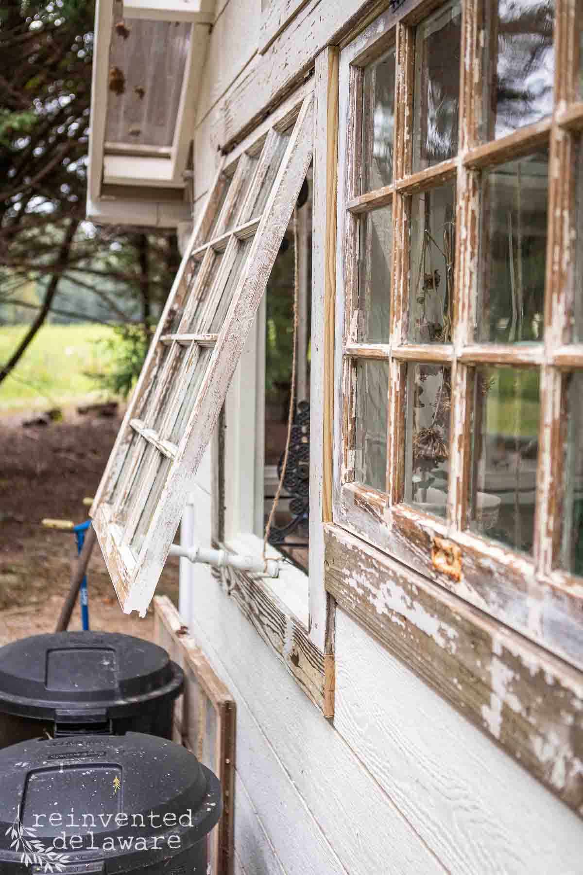 A weathered, white-painted wooden garden shed with two large, old-fashioned windows. One window is propped open. Below the window, there are two black plastic trash cans used for composting. The she shed is surrounded by trees and greenery. An outdoor hose is visible on the wall by the trash cans.