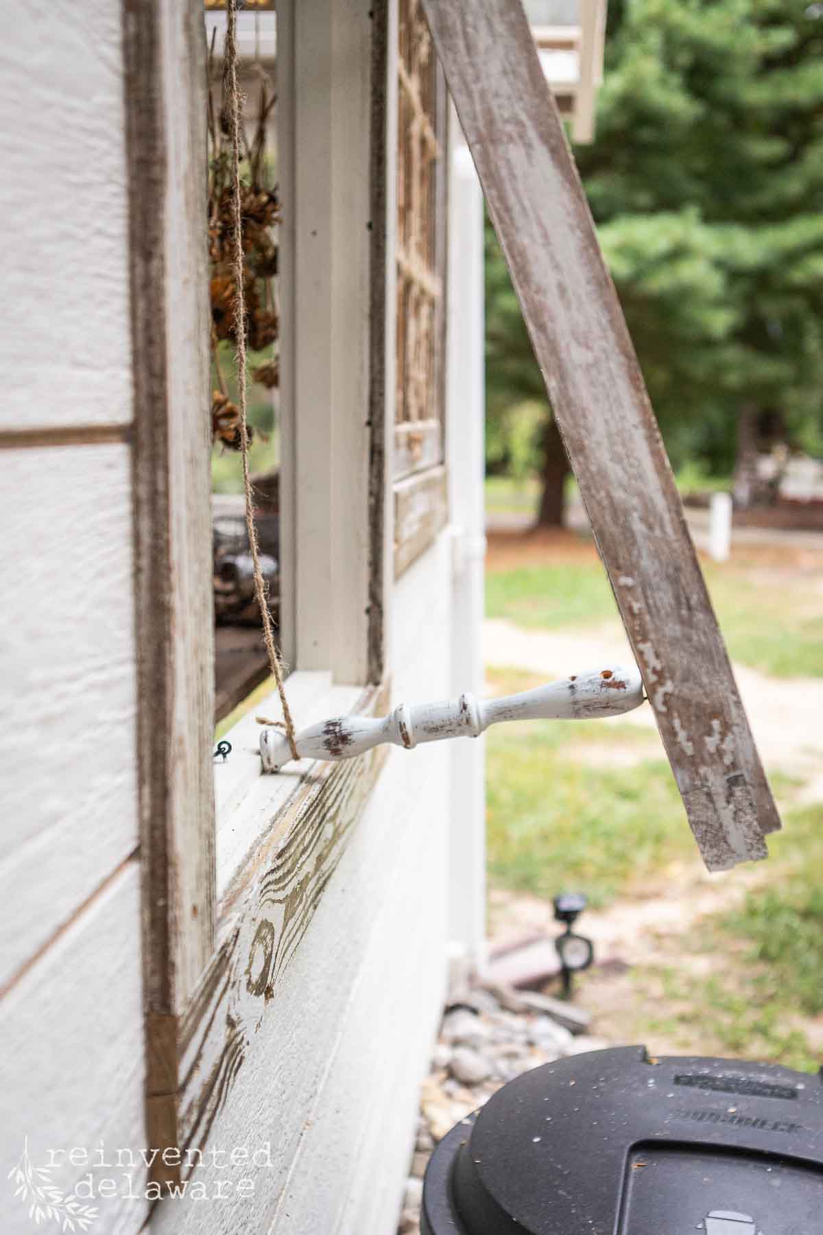 A close-up of a weathered wooden shutter held open by a prop against the side of a white-painted garden shed. The window frame and siding show signs of wear. In the background, a green forest and a path are visible. The text "reinvented delaware" is in the bottom-left corner.
