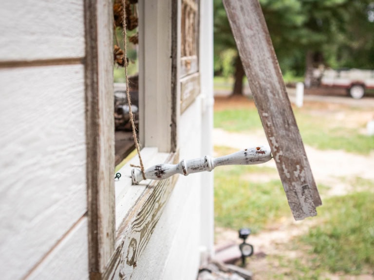 Close-up of a weathered, white-painted wooden window shutter, slightly open with a broken hinge. The background shows a blurred green outdoor space with trees and an off-white pickup truck in the distance.