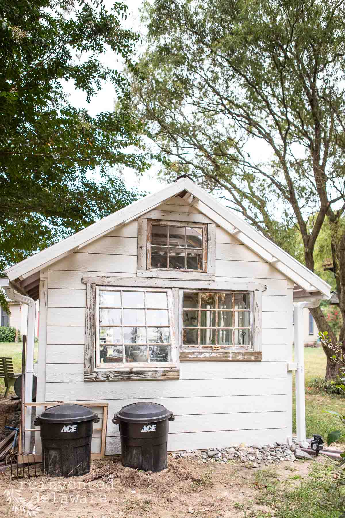 A small white wooden shed with two large windows and one smaller window above. Two black ACE trash bins are in front of the shed. Trees surround the area, and a glimpse of another building is visible to the left. The setting looks like a yard or garden.