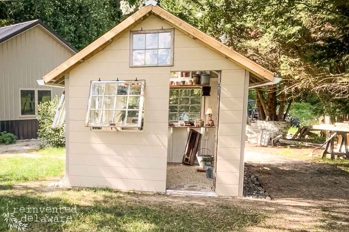A beige shed with a partially open window displaying a small garden shed inside. The shed has a window on the roof peak and is surrounded by grass and trees. Some tools and materials are visible inside. A wooden structure is partially visible in the background.