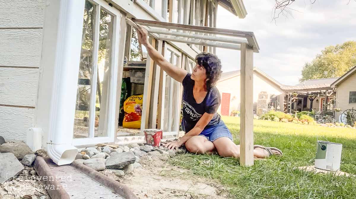 A person crouches on the ground outside, painting a white trim of a window on a small building. They are wearing a black t-shirt and blue shorts. Various tools and paint supplies are scattered around. There are plants and other buildings in the background.