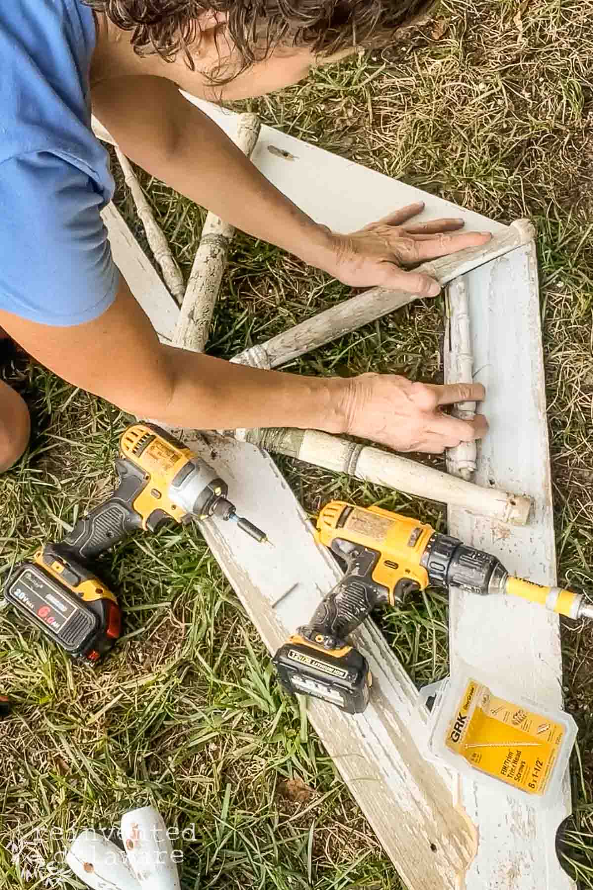A person in a blue shirt is working on a wooden frame outdoors, using a power drill and other tools. Various tools and supplies, including a second power drill and a tube of adhesive, are scattered on the grass around them.