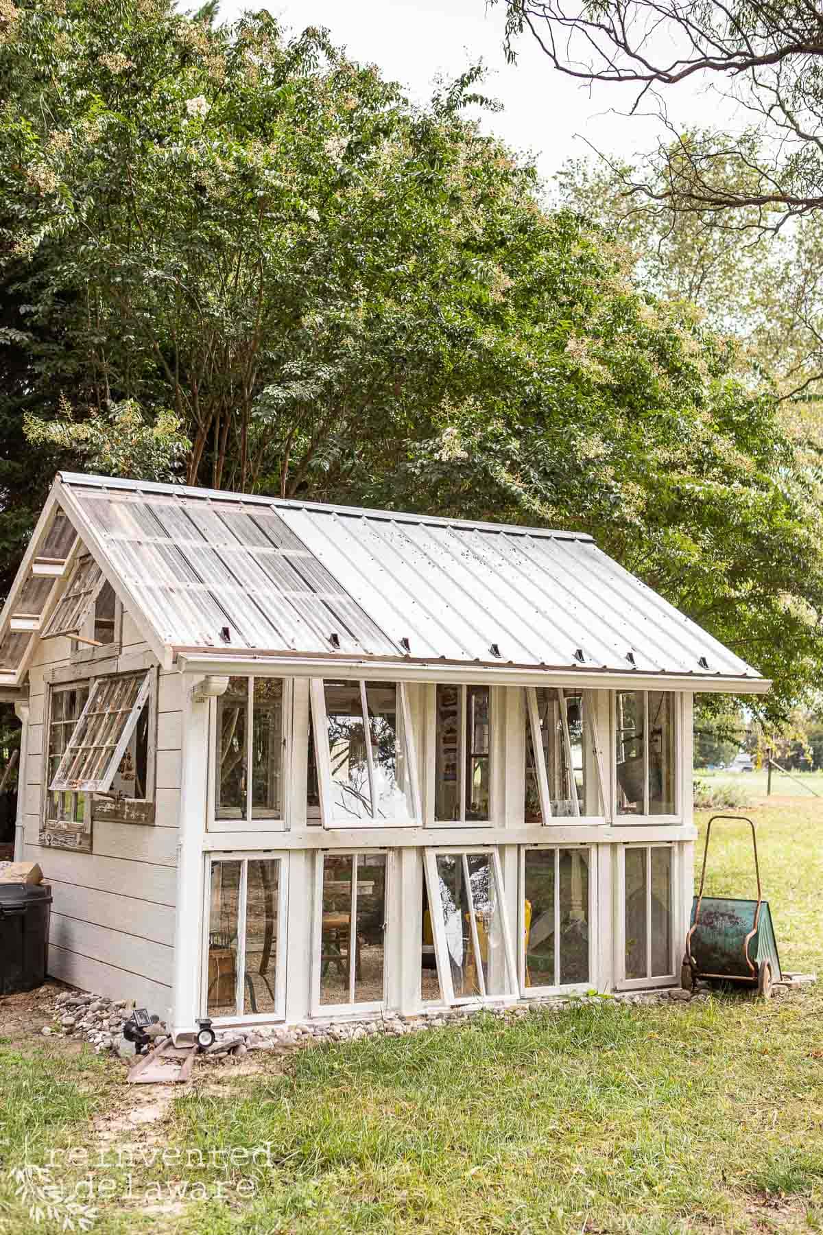 A small greenhouse constructed from repurposed glass windows and doors, surrounded by lush green trees. The white-framed structure features a pitched roof and several windows for ventilation. A wheelbarrow and garden tools are visible beside the greenhouse.
