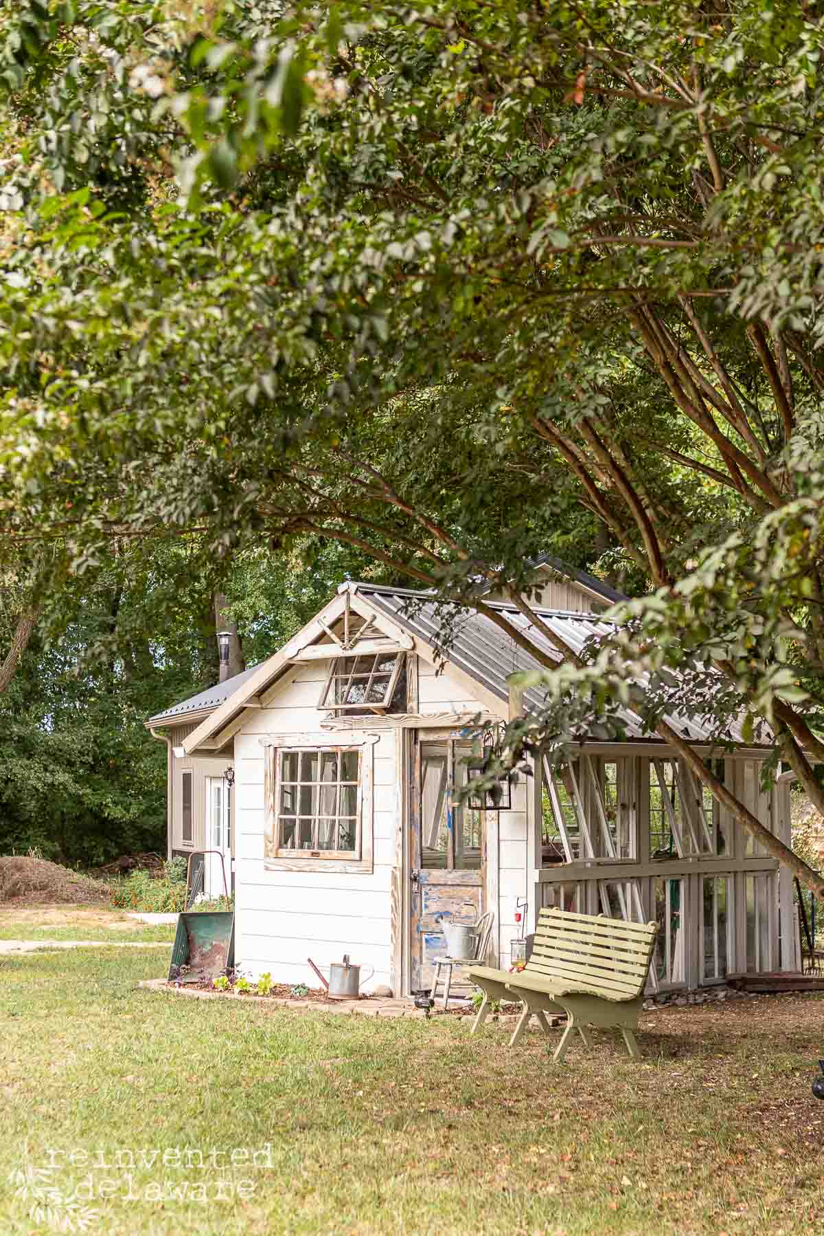 A small, rustic white shed is surrounded by lush greenery and trees. It features large windows and a metallic roof. In front, there's a wooden bench and some gardening tools. The overall setting is peaceful and inviting.