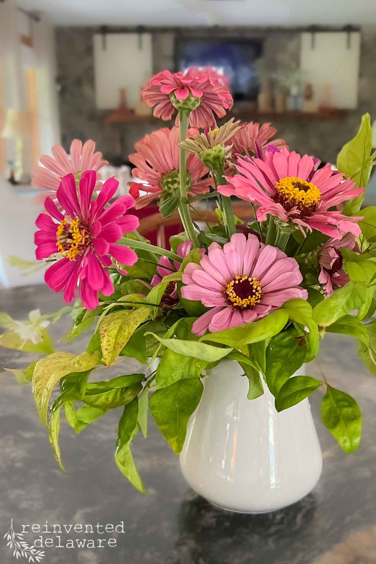 A white ironstone pitcher filled with vibrant pink and magenta zinnias and green foliage sits on a dark countertop. The out-of-focus background features various home decor elements, providing a cozy and inviting ambiance.