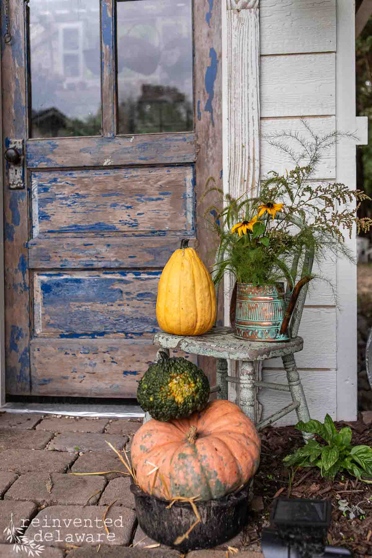 A rustic, weathered door with peeling blue paint is decorated with a tall yellow gourd. Beside the door is a small wooden chair holding a potted plant with yellow flowers. In front, two pumpkins, one green and one orange, are stacked on the ground.