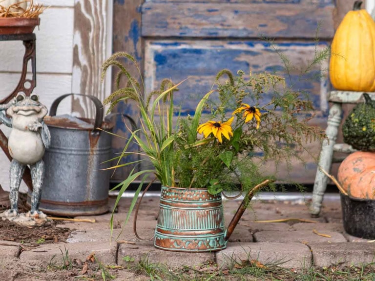 A rustic scene with a weathered blue door in the background. In the foreground, there's a decorative metal watering can holding assorted fresh flowers and greenery. Nearby, a frog statue, a pumpkin, and a small chair add to the charming outdoor display.