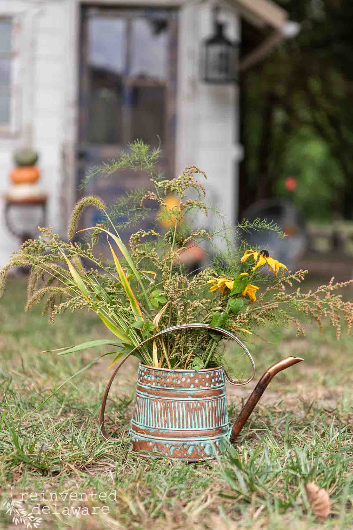 A rustic watering can repurposed as a planter holds a vibrant assortment of wildflowers and greenery. The scene is set outdoors on a grassy area with a blurred background featuring a small white building adorned with pumpkins and a blurred hanging lantern.