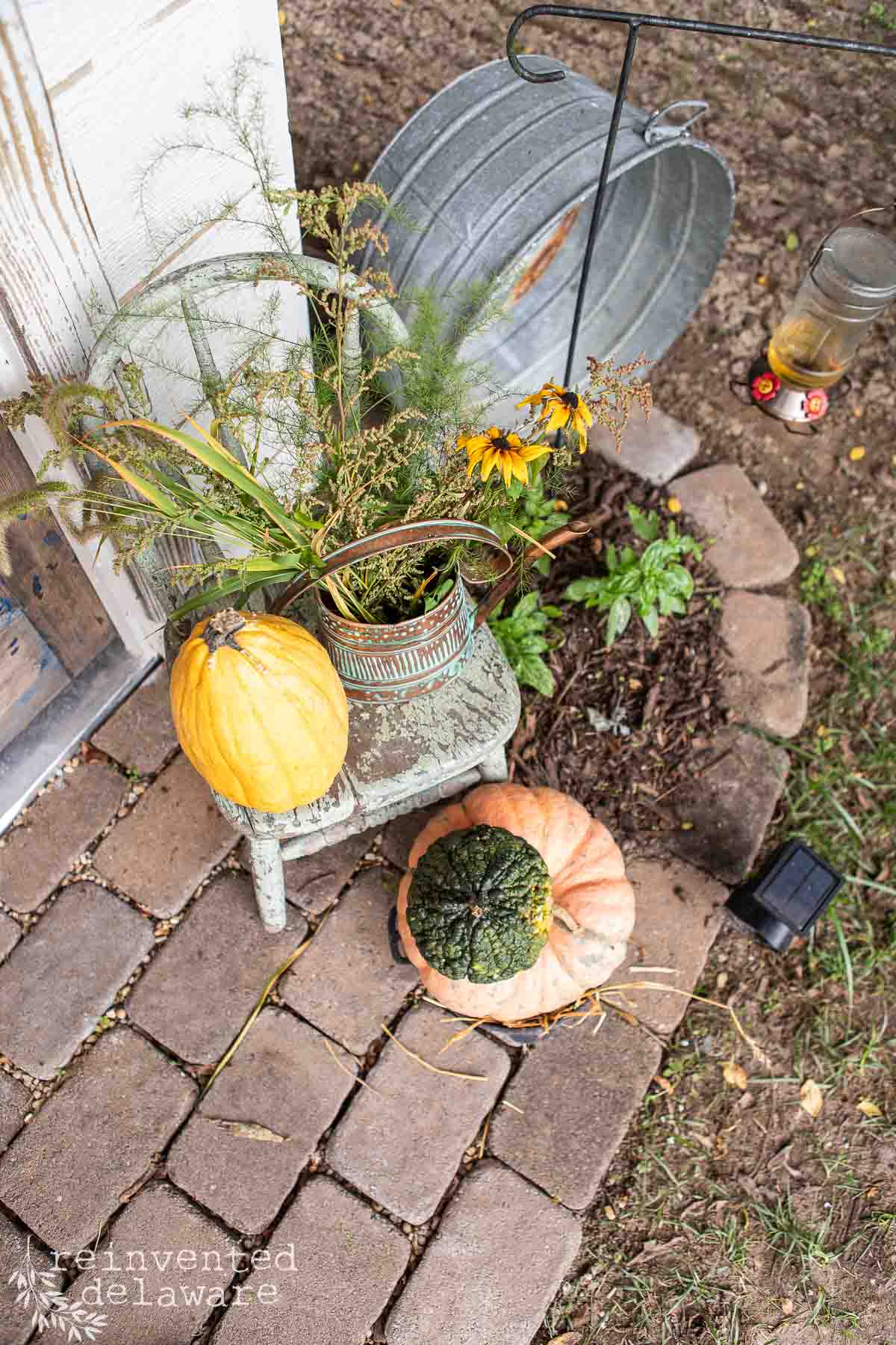 A rustic outdoor scene with a yellow gourd and a green-and-orange squash on brick pavers. There's a decorative metal watering can with plants, a galvanized metal tub, and a bird feeder hanging from a metal hook. Grass and dirt can be seen in the background.