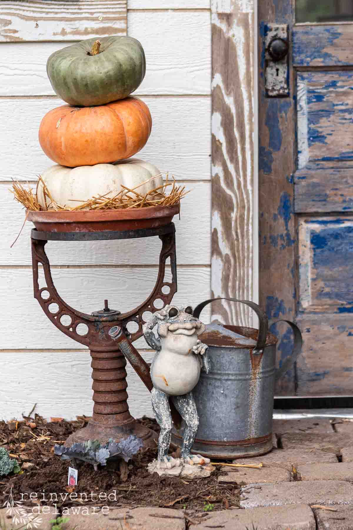 A whimsical porch display features a stack of green, orange, and white pumpkins on a vintage cast iron water tank stand. A weathered metal watering can and a charming frog statue holding binoculars sit beside the stand. The background includes a rustic, worn blue and white wooden door.