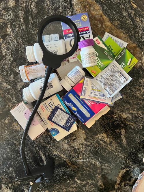 A pile of various medication bottles and boxes scattered on a marble surface, with a magnifying glass positioned overhead, highlighting some of the text on the packaging.