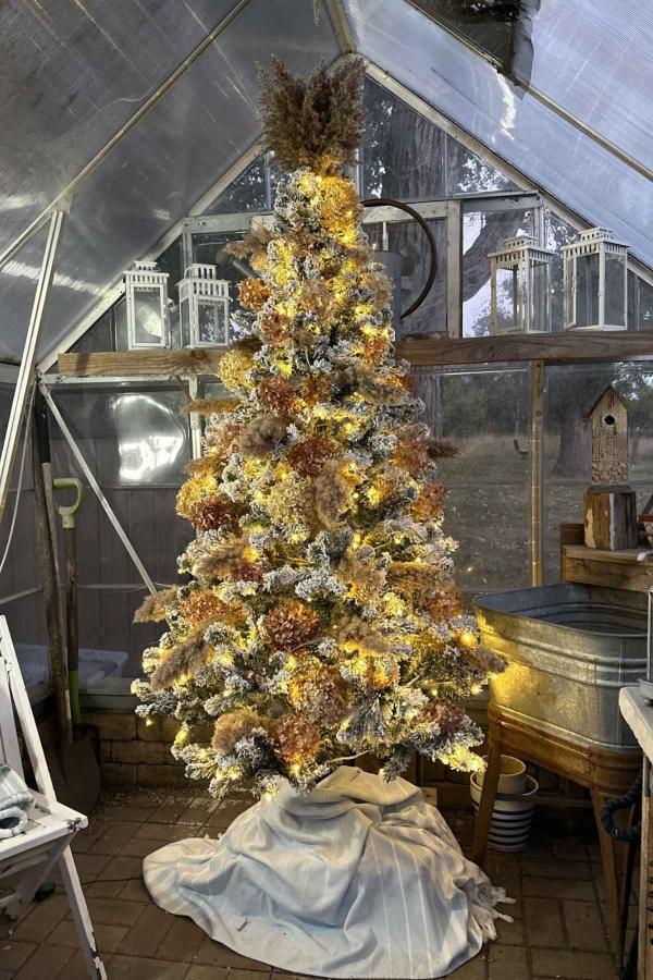 A decorated Christmas tree stands in a greenhouse, adorned with lights and festive ornaments. The tree is placed on a white cloth, surrounded by rustic decor, lanterns, and gardening tools. The setting has a cozy, rustic ambiance.