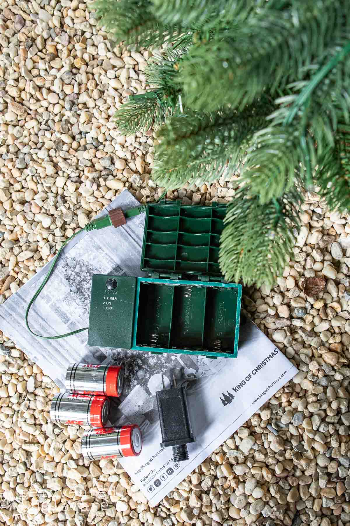 A green electronic device with open compartments lies on a newspaper atop pebbles. Nearby, there are three red and silver D batteries and a section of artificial pine tree branches.