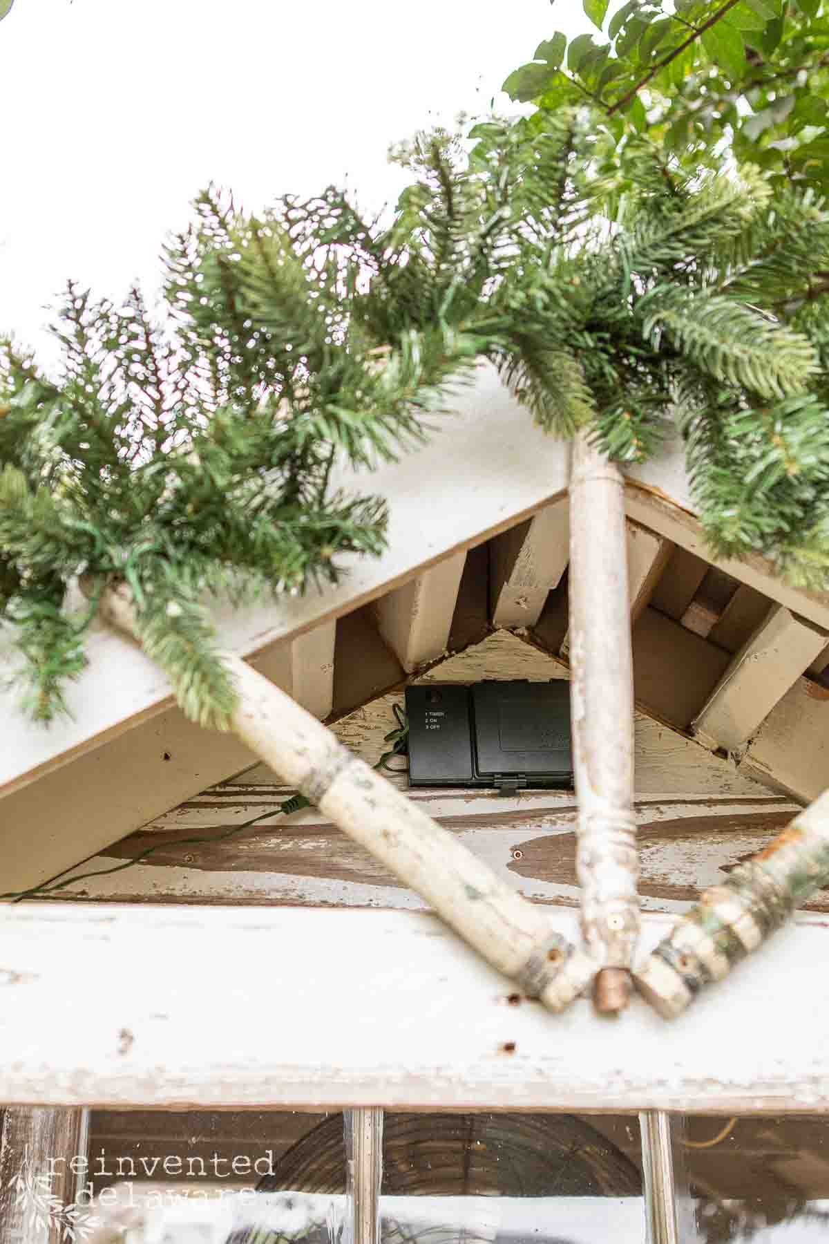 Close-up of a wooden she shed with two rustic, white-painted spindles forming an 'X' design. Evergreen branches are draped over the top, and part of a vintage glass window is visible below.