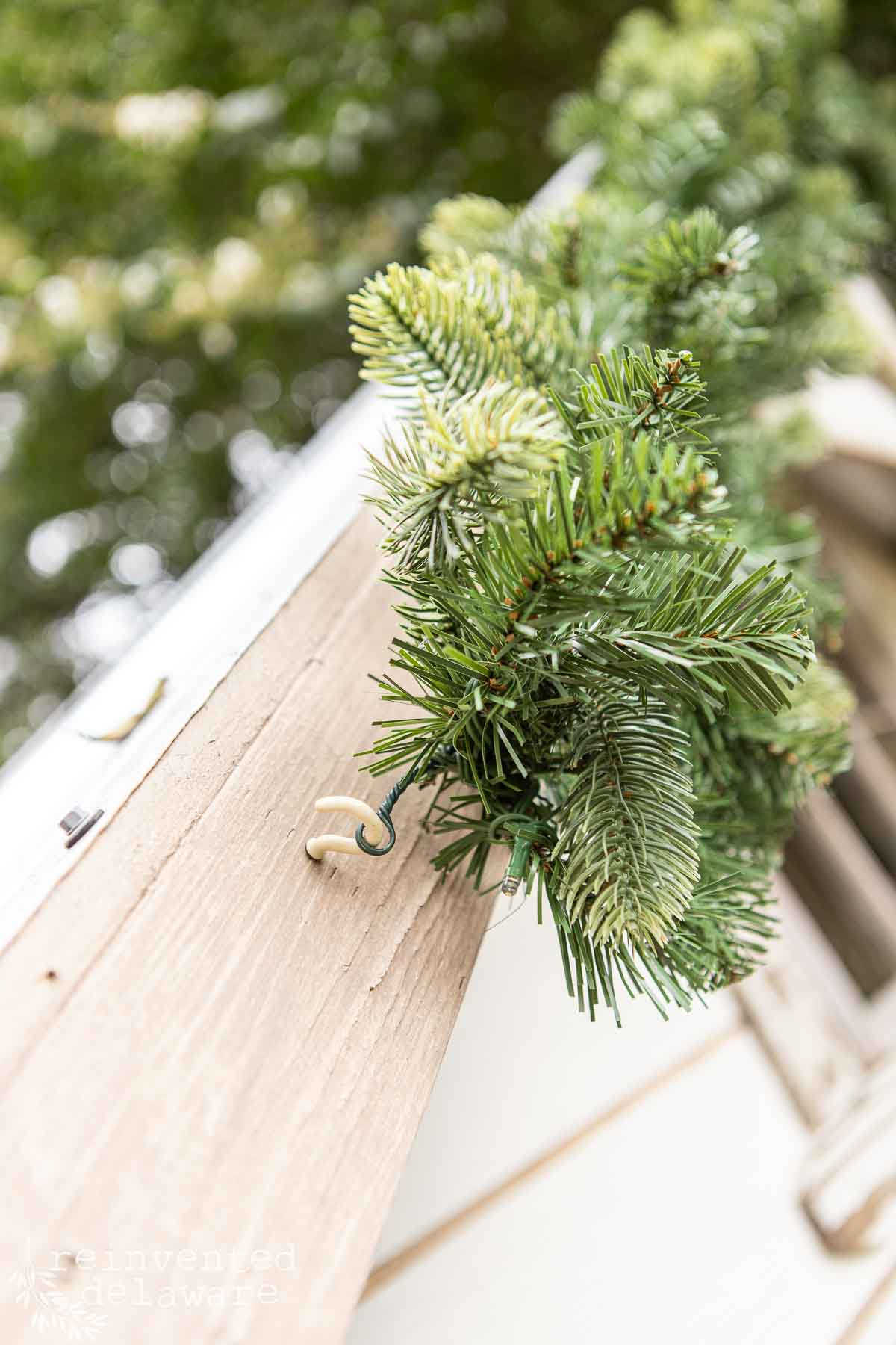Close-up of a green fir garland hanging on the corner of a wooden structure. The garland is secured by a hook, and the image has a soft, blurred background of greenery.