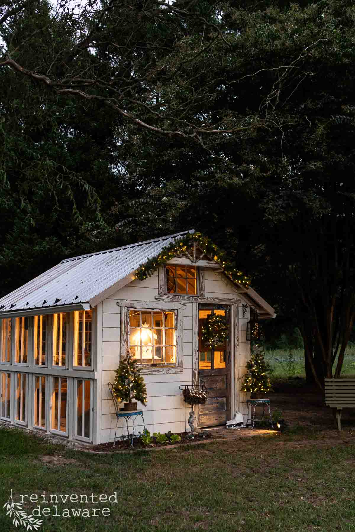 A cozy, illuminated greenhouse with a white exterior decorated with string lights and greenery. It sits nestled among dark trees, creating a warm, inviting contrast at twilight. There's a bench on the right side.