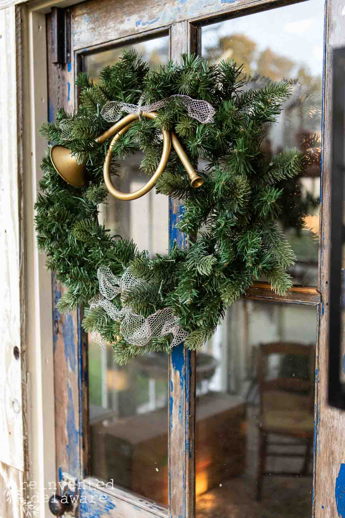 A festive green wreath with a decorative horn and mesh ribbon hangs on a rustic, weathered wooden door. The reflection of a chair is visible in the glass panel behind the wreath.