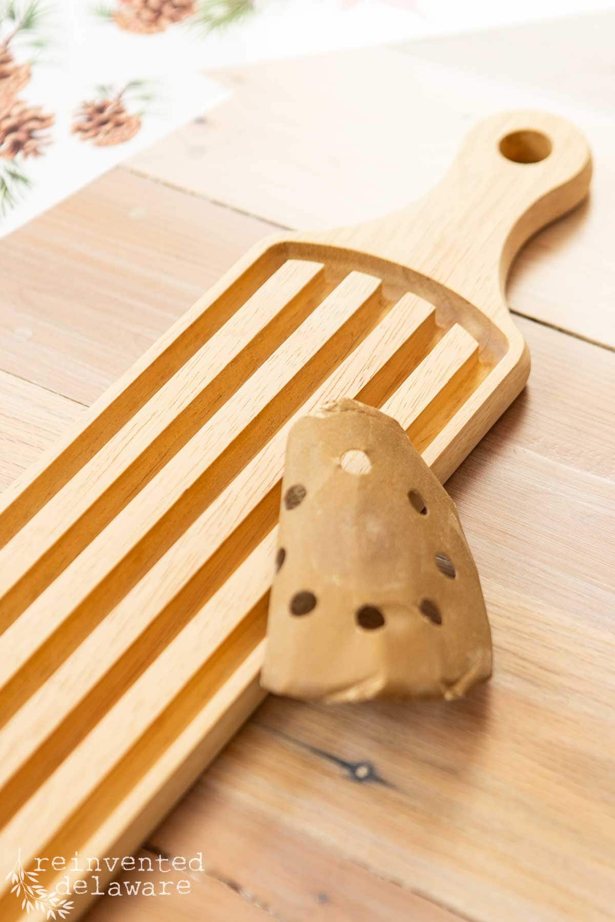 A wooden breadboard with grooved design rests on a light wood surface. A piece of sandpaper with holes is placed partially over it. Pinecone and foliage patterns are visible in the background.