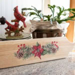 A festive wooden cutting board with poinsettia and pinecone transfer design rests on a kitchen countertop. Nearby, a decorative figurine of two red cardinals sits next to a lush green plant in a basket.