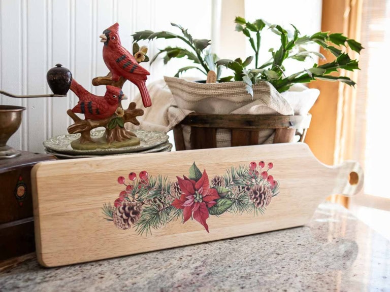 A festive wooden cutting board with poinsettia and pinecone transfer design rests on a kitchen countertop. Nearby, a decorative figurine of two red cardinals sits next to a lush green plant in a basket.