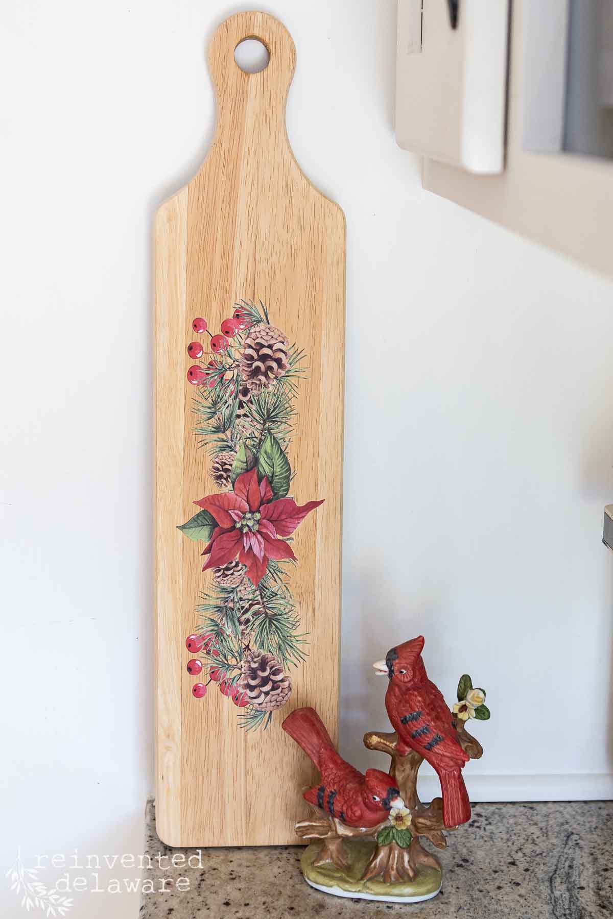 A wooden cutting board with a festive design of poinsettias, pinecones, and greenery. Next to it is a decorative ceramic figure of two red cardinals perched on a small branch.