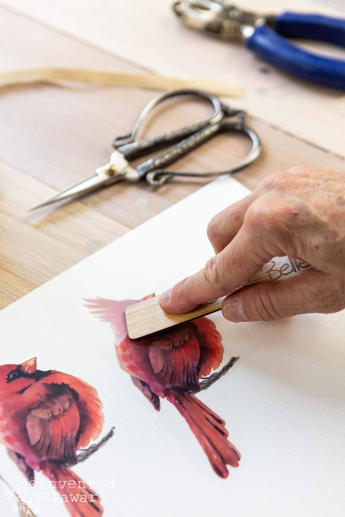 A hand is burnishing a watercolor image of a red cardinal on paper using a small wooden tool. Scissors and a pair of blue pliers are in the background on a wooden surface.