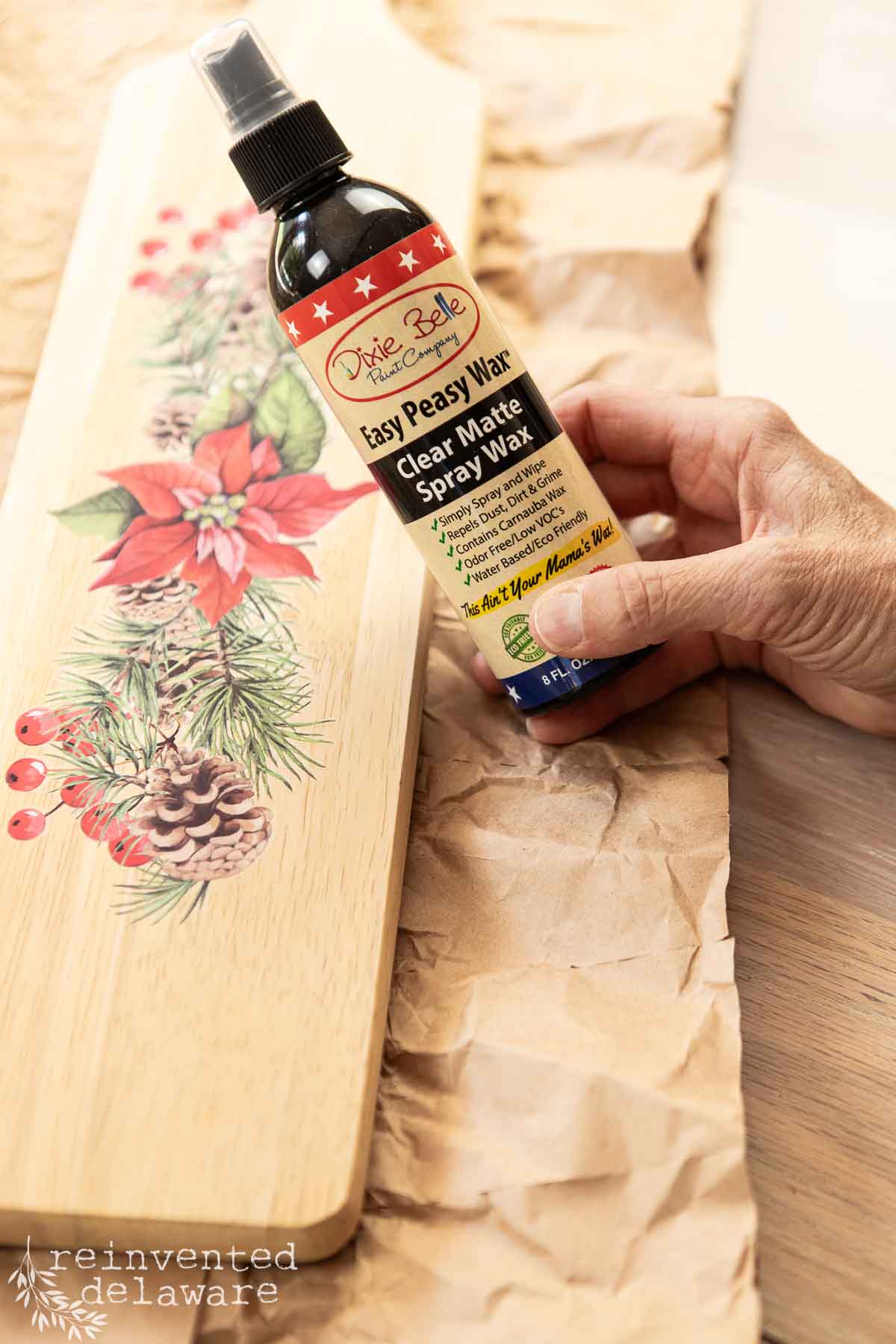 A hand holding a bottle of "Dixie Belle Easy Peasy Wax" spray next to a wooden cutting board decorated with a painted poinsettia and pinecones. The board rests on crumpled brown paper.