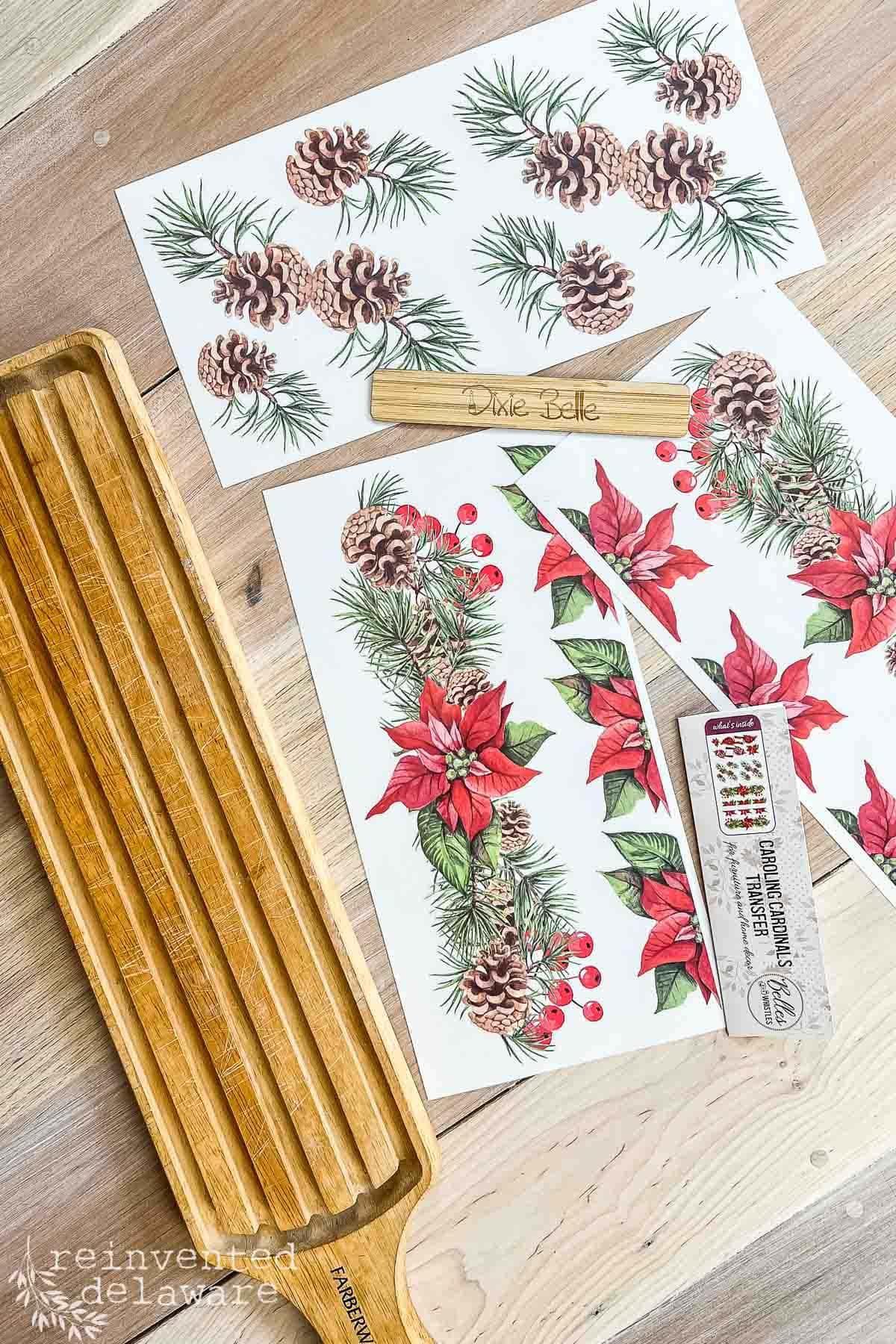 A wooden cutting board with carved grooves sits beside sheets of decorative Christmas-themed transfers featuring pinecones, poinsettias, holly, and berries. A small wooden stick labeled "Dixie Belle" and a small instruction paper are also visible.