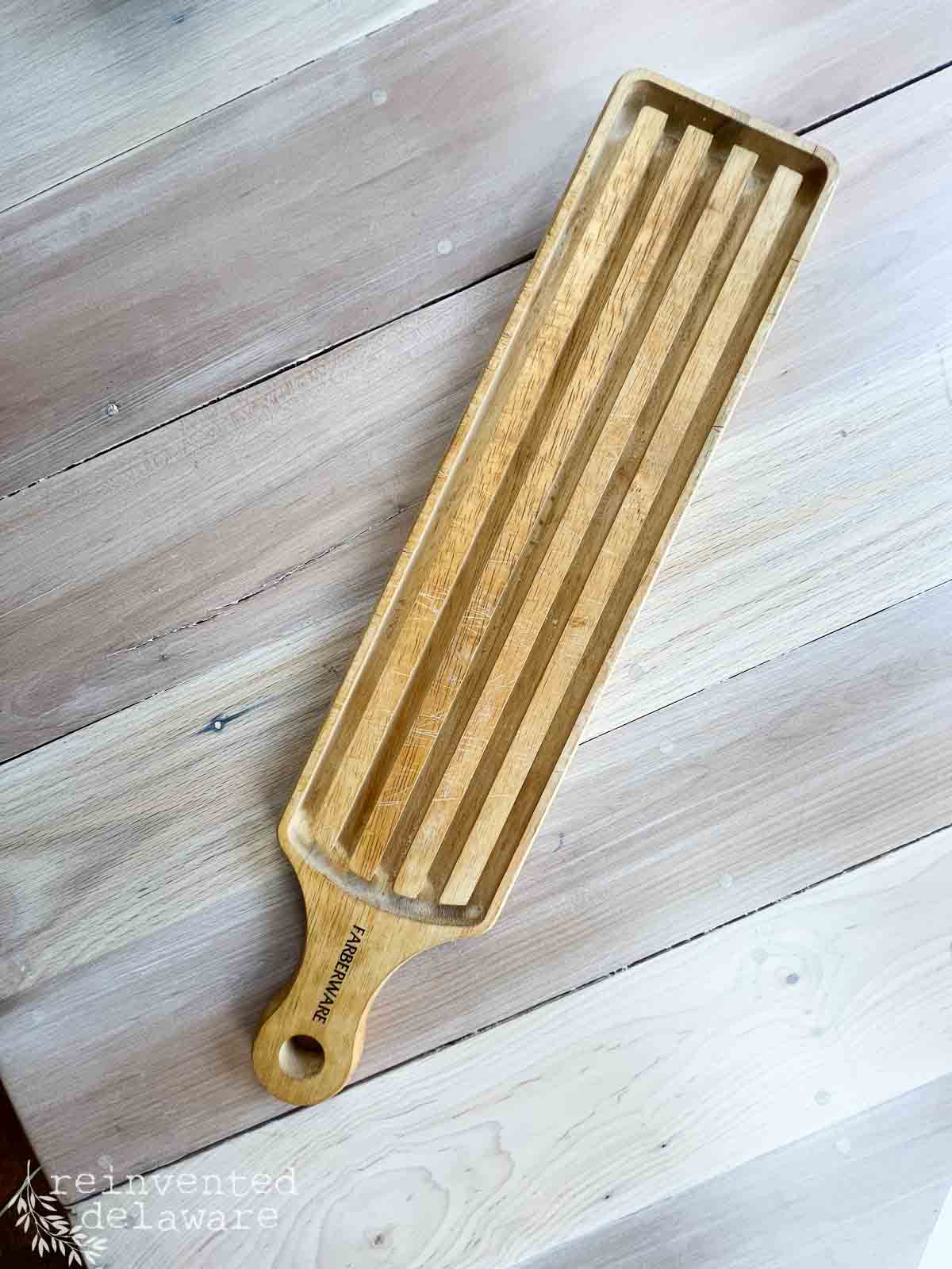 A long, rectangular wooden breadboard tray with grooves and a handle rests on a light wood surface. The handle is engraved with "Farberware"