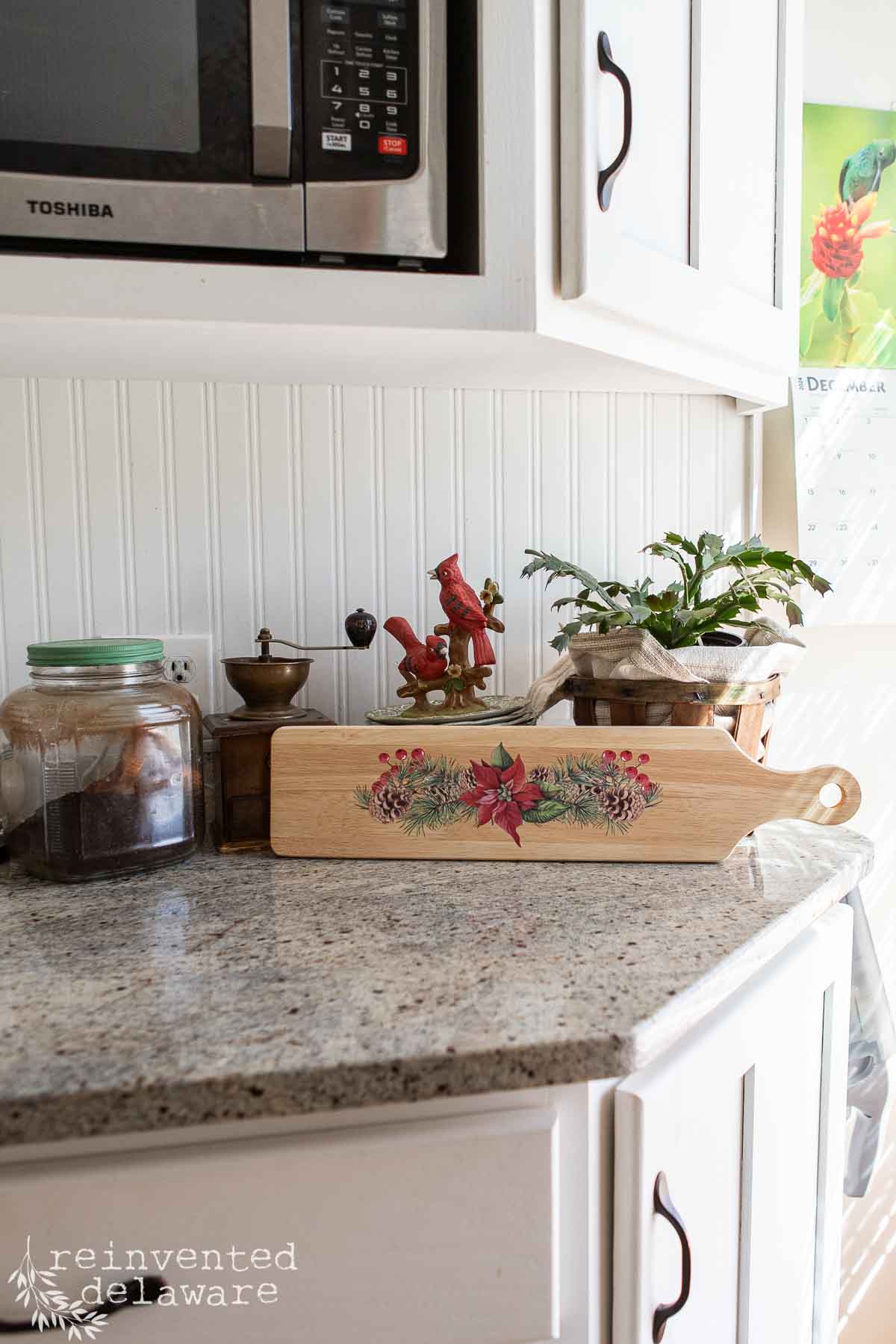 Cozy kitchen with a decorative cutting board featuring poinsettias. Nearby are a vintage coffee grinder, a jar of coffee beans, and a potted plant on a speckled countertop under a wall-mounted microwave.