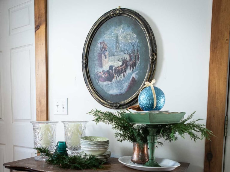 A wall showcases an oval winter scene painting of a sleigh and reindeer. Below, a table holds a green pedestal with a blue ornament, glassware, plates, and evergreen branches, creating moody Christmas decor that's both elegant and inviting.