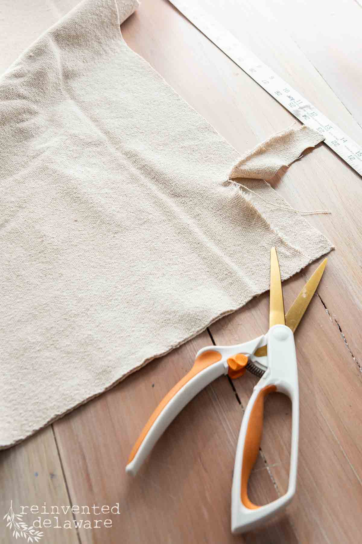 A pair of fabric scissors is resting on a wooden table alongside a piece of light beige material. A measuring tape is also visible beside the fabric.