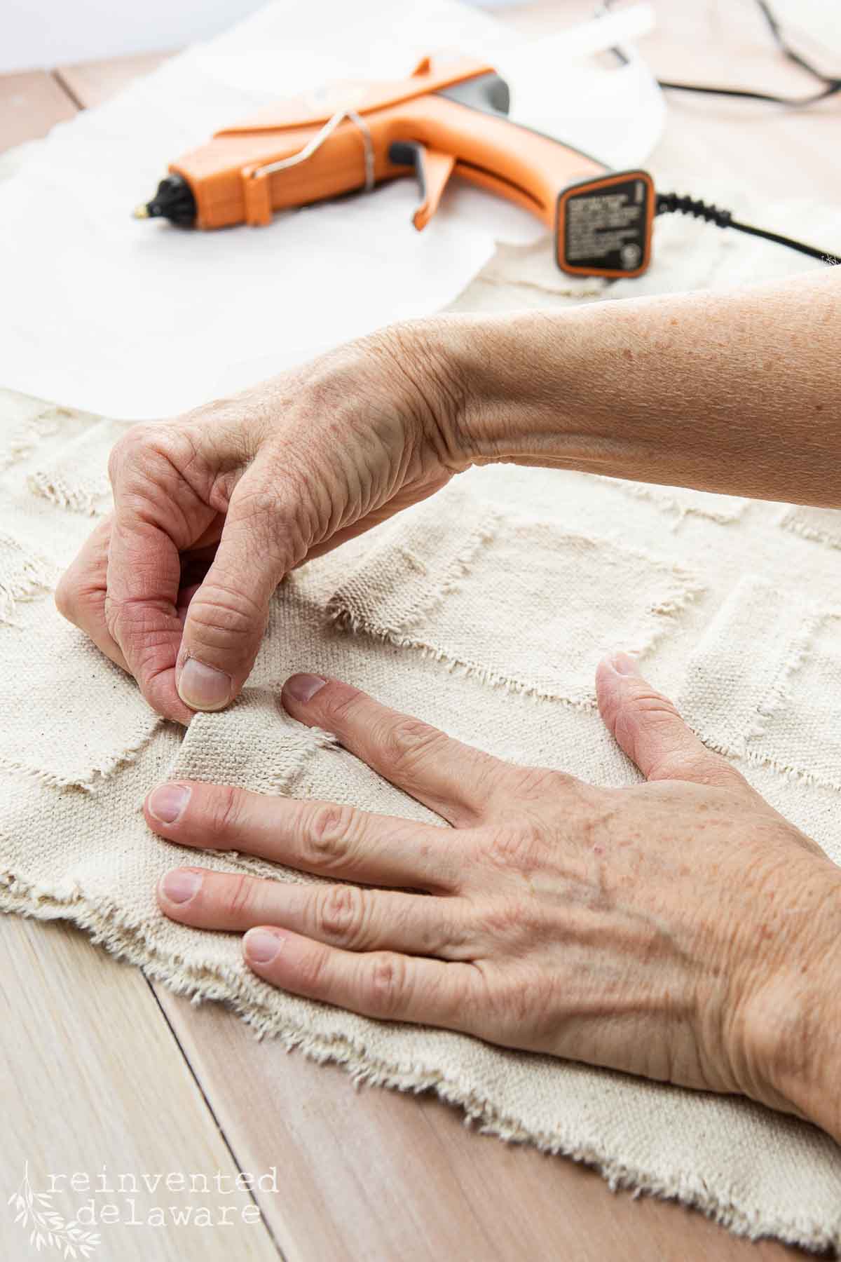 Close-up of hands crafting with burlap on a wooden table. A glue gun rests nearby. The person is arranging cut pieces of burlap, displaying a project in progress. The image has a watermark, "reinvented delaware," in the lower-left corner.