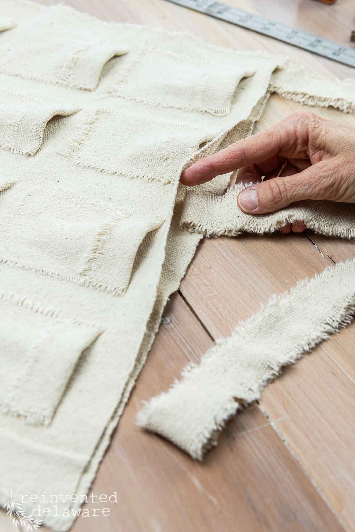 A hand points to a piece of beige fabric with square patterns and frayed edges laid on a wooden surface. A frayed fabric strip is beside the main piece. A measuring tape is partially visible in the background.