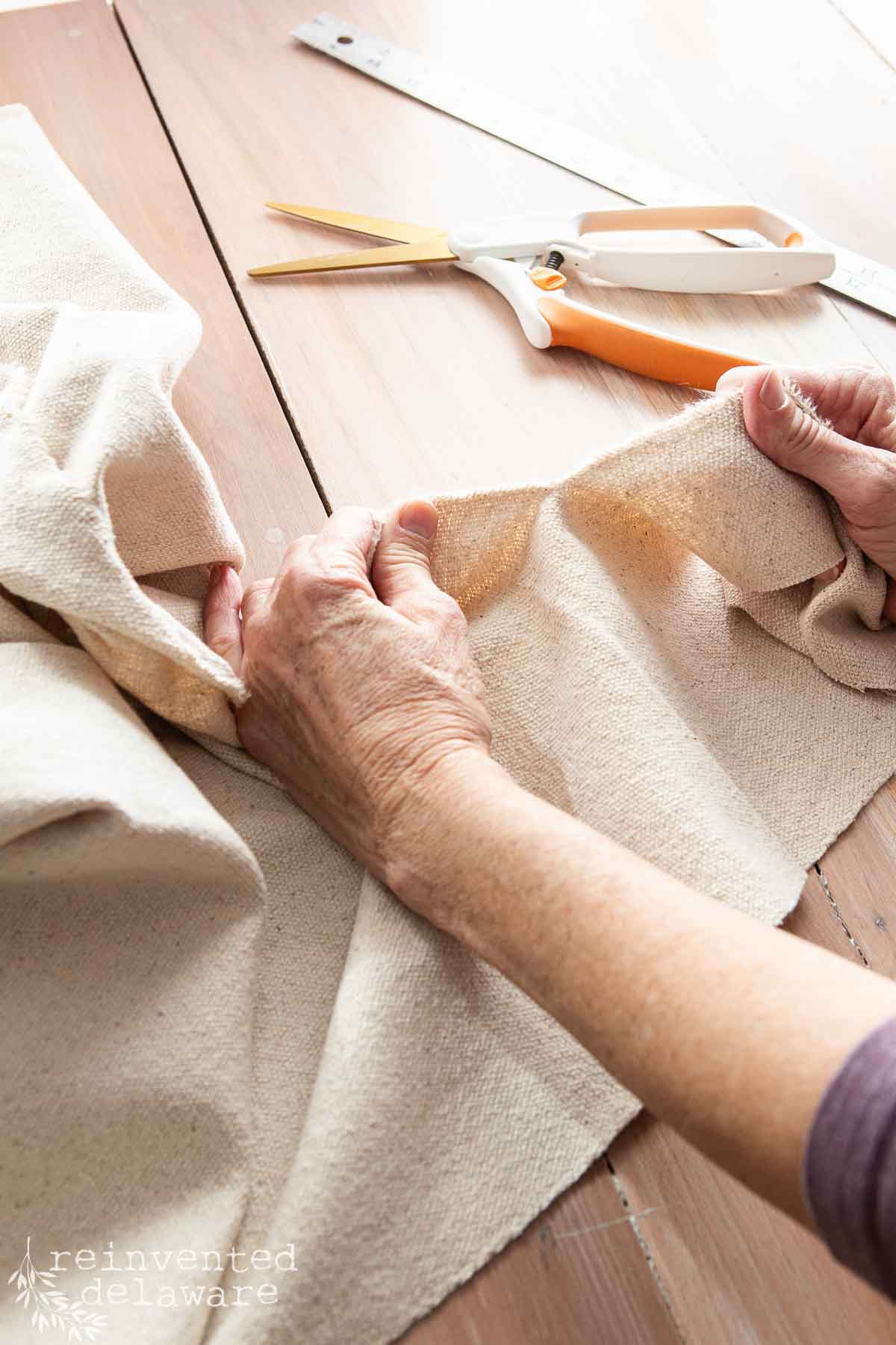 Close-up of hands folding a piece of beige grainsack fabric on a wooden surface, with a pair of orange-handled scissors and a metal ruler in the background.