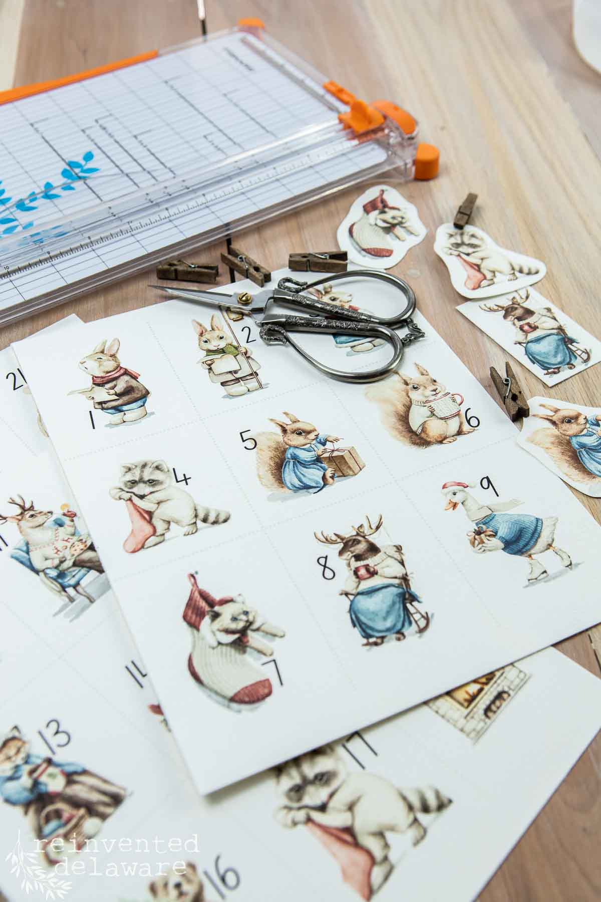 A table with a paper cutter, scissors, and paper sheets featuring vintage animal illustrations numbered 1 to 9. Some animals are dressed in clothes and carrying sacks. Small clips are scattered around. Pieces used for a DIY advent calendar.