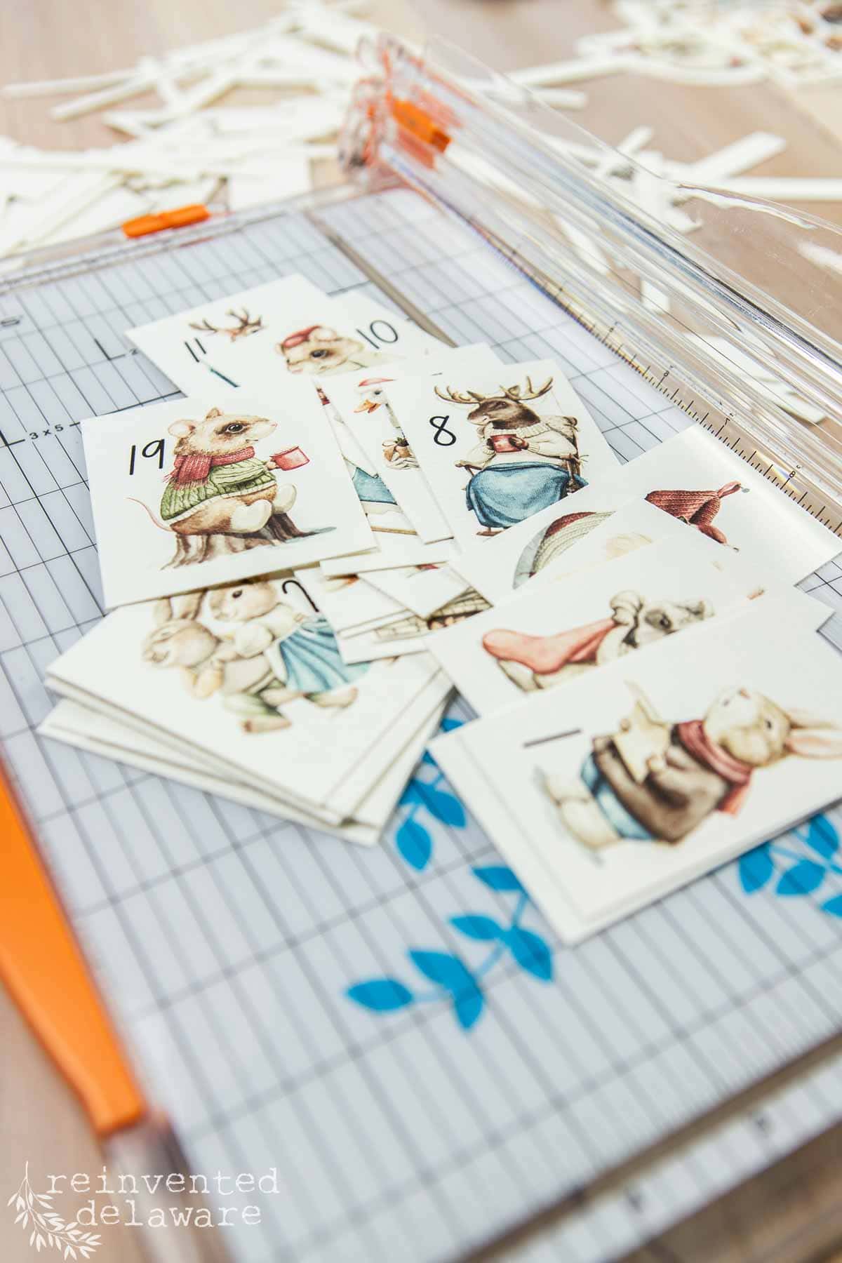 A stack of paper cards featuring illustrations of animals dressed in clothes is being cut using a paper trimmer with an orange edge. Perfect for creating a DIY advent calendar, the scene is set on a grid cutting mat with small paper scraps scattered around.
