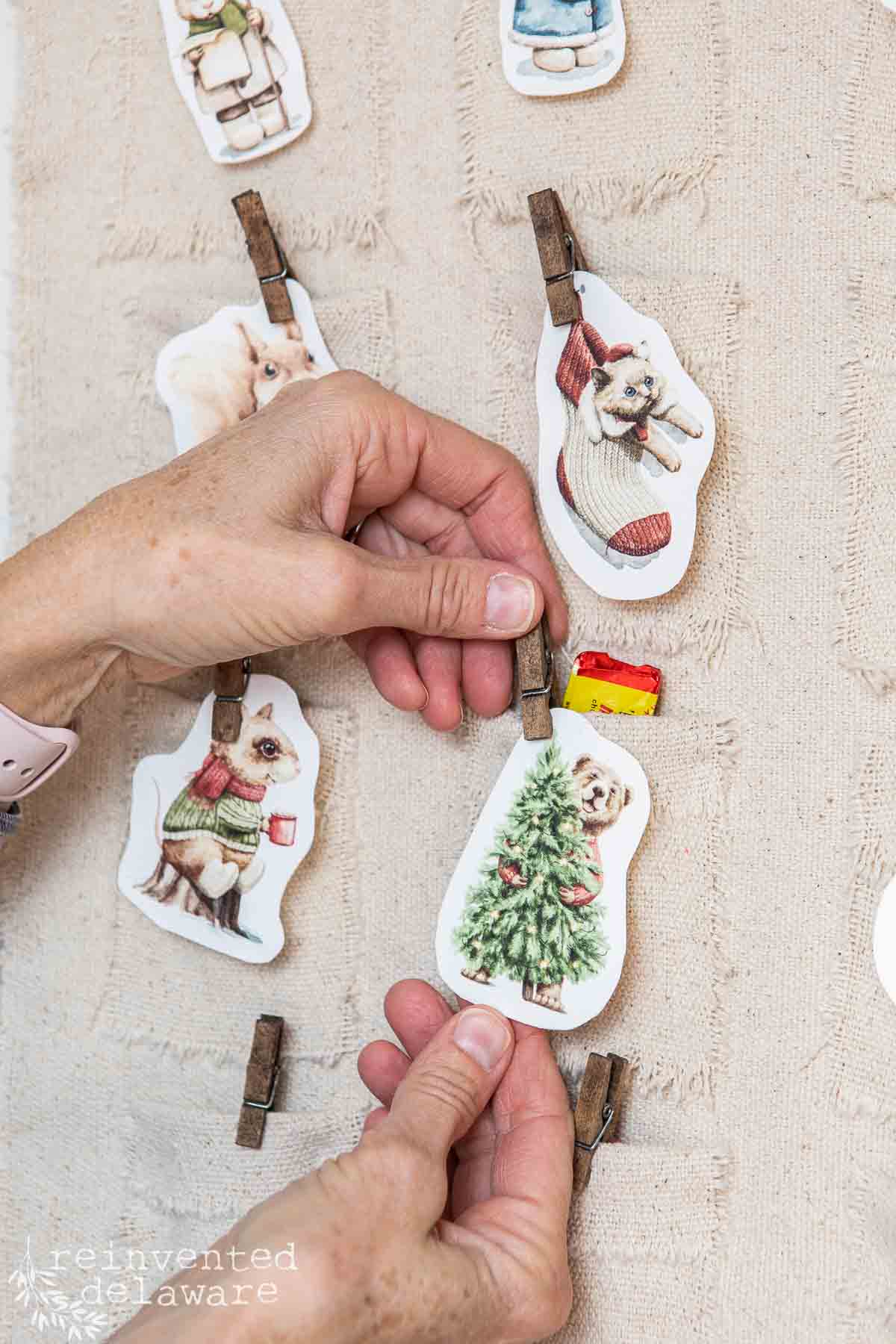 Hands are attaching small, vintage-style animal illustrations to a textured fabric background using mini clothespins, creating a charming DIY advent calendar. Each animal is dressed in winter attire, holding festive items like presents and trees.