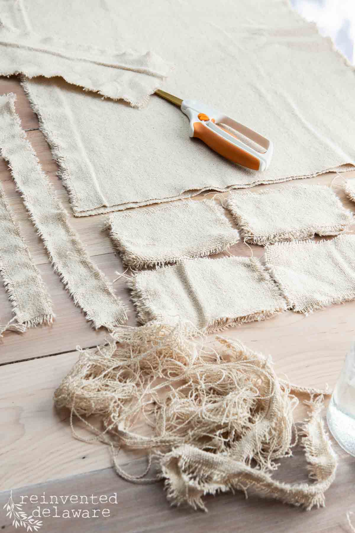 A table with frayed fabric pieces and a pair of orange-handled scissors. The cloth is arranged in rectangles and strips. Loose threads are scattered around.