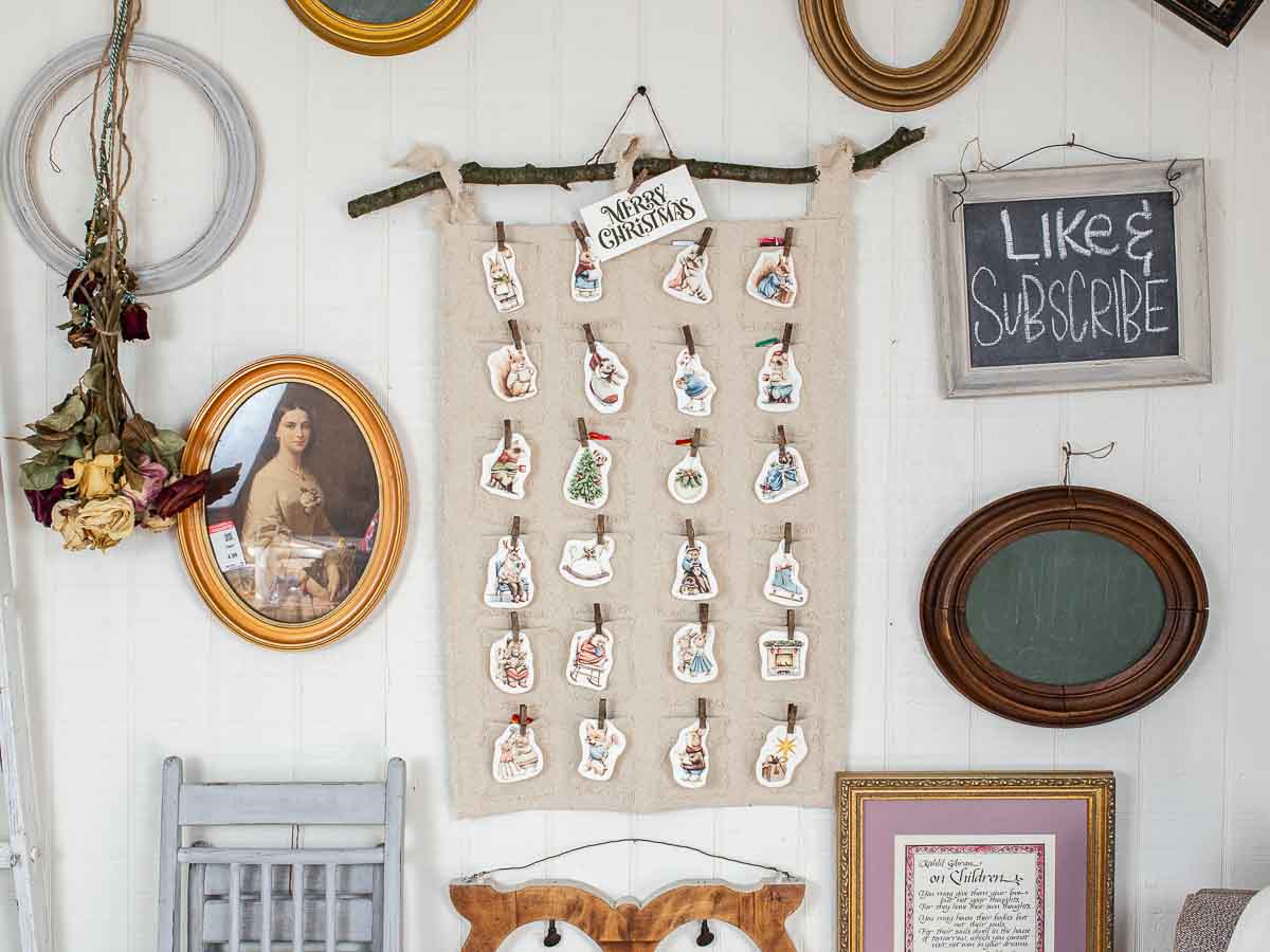 A wall adorned with a charming DIY Christmas advent calendar, showcasing small wooden cutouts. Nestled among frames, dried flowers, and a "Like & Subscribe" sign, the calendar hangs gracefully from a branch with a festive "Merry Christmas" tag.
