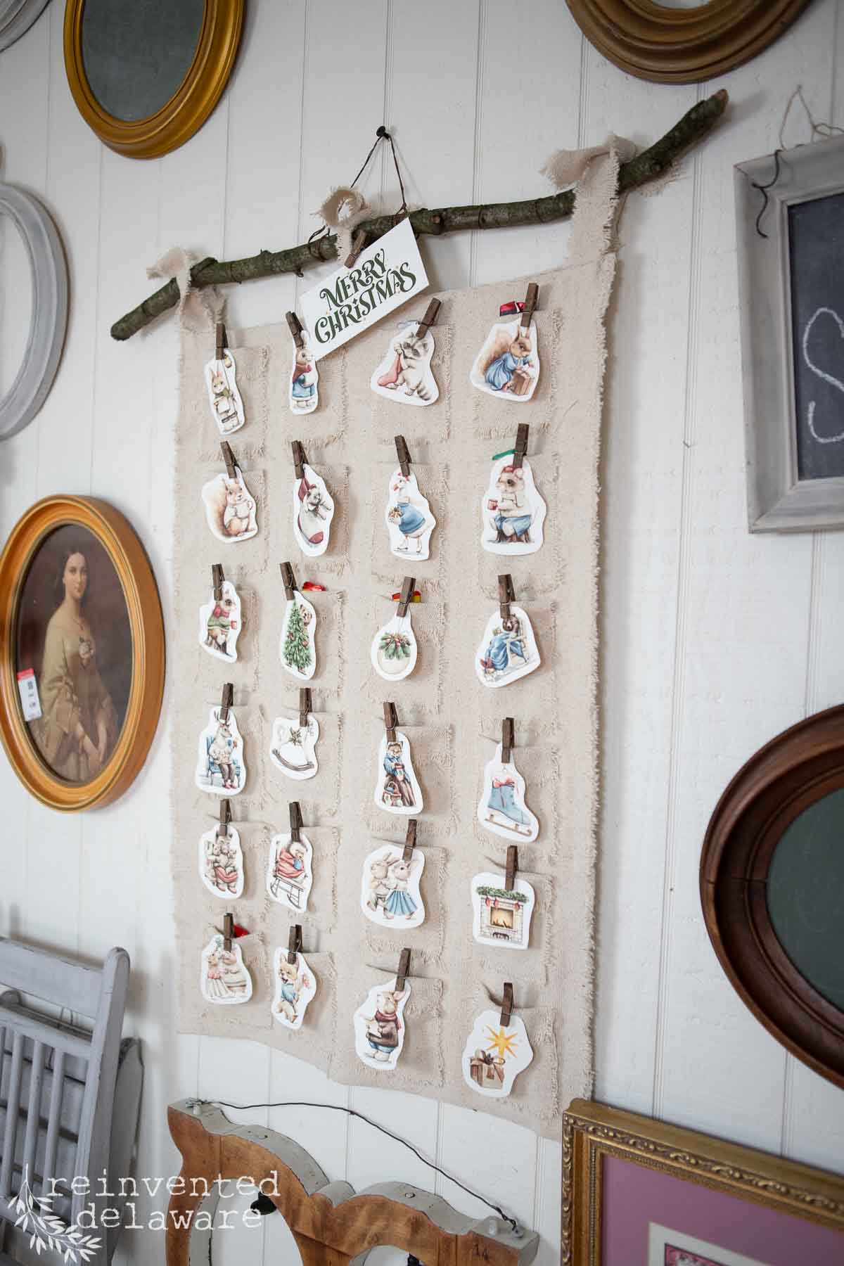 A DIY advent calendar made from a branch and fabric showcases small, illustrated cards clipped with mini clothespins. A "Merry Christmas" sign tops the display, surrounded by vintage picture frames on a white wall, creating a charming festive focal point.