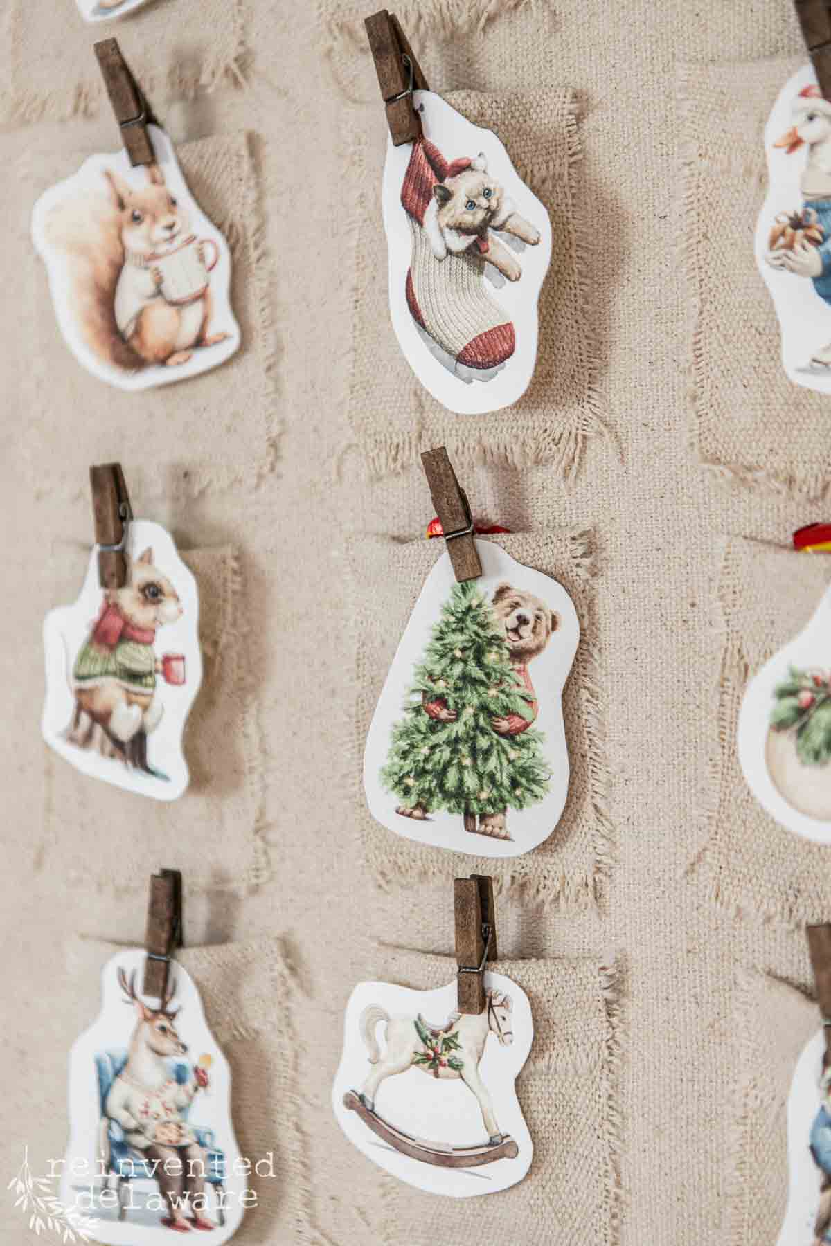 Illustrated Christmas cards featuring woodland creatures hanging on a textured fabric with clothespins evoke the charm of a DIY advent calendar. Some images include a cat in a stocking, a dog with its Christmas tree, and a playful rocking horse.