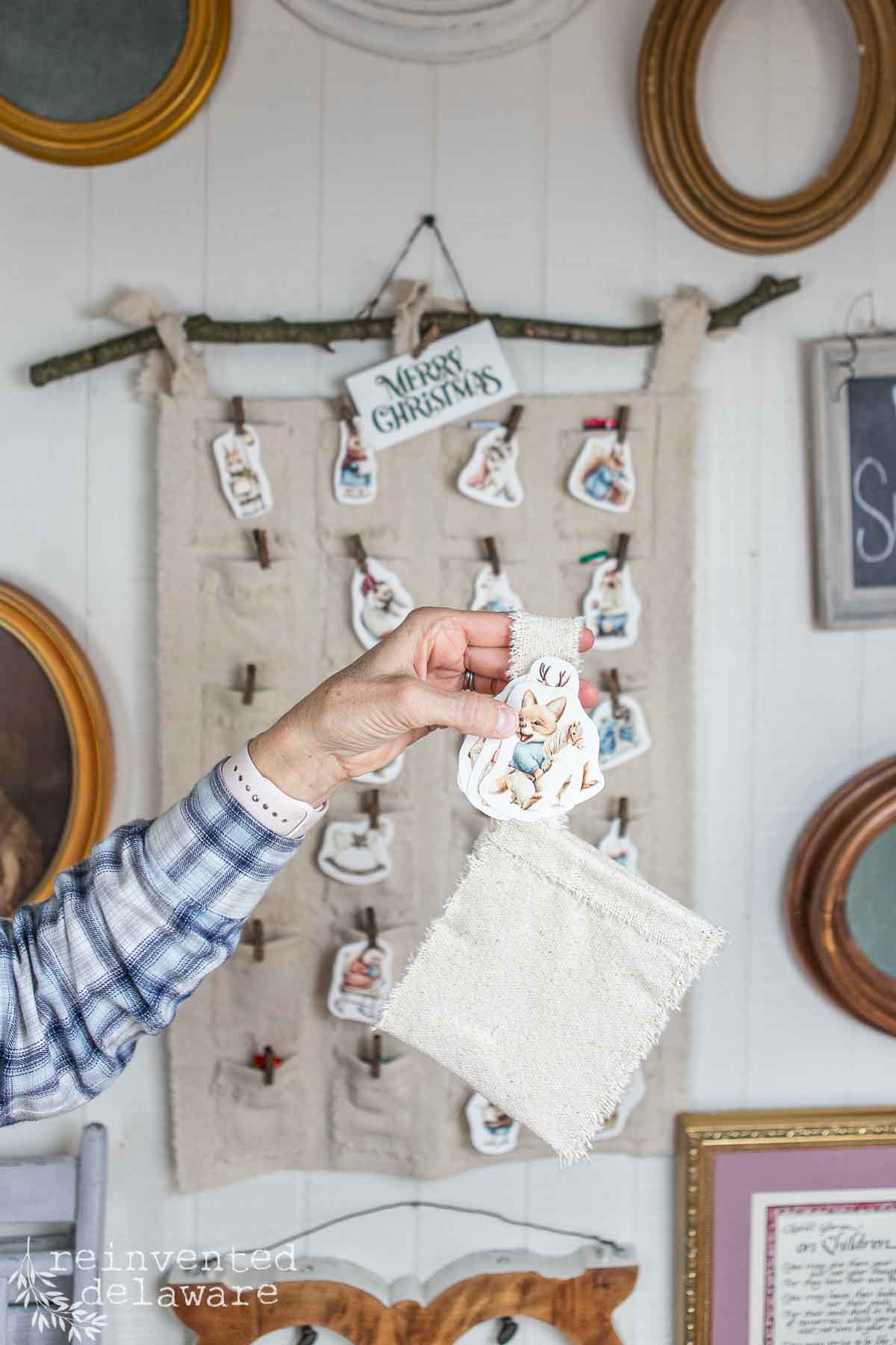 A person holds a fabric square bag with a vintage-style woodland creature card. Behind are more cards hanging on a grain sack fabric diy advent calendar with "Merry Christmas" at the top. Surrounding the display are framed pictures and mirrors on the wall.