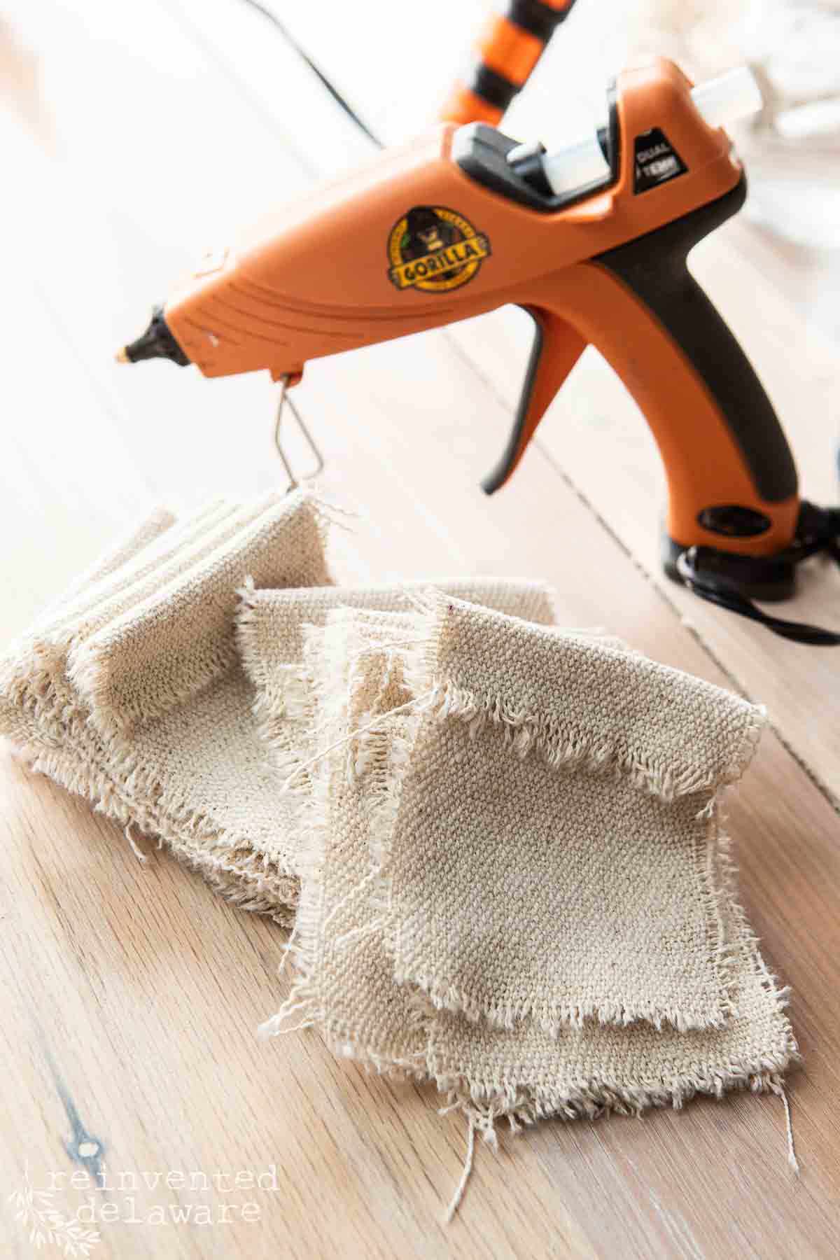 A stack of frayed fabric squares next to an orange hot glue gun on a wooden surface.