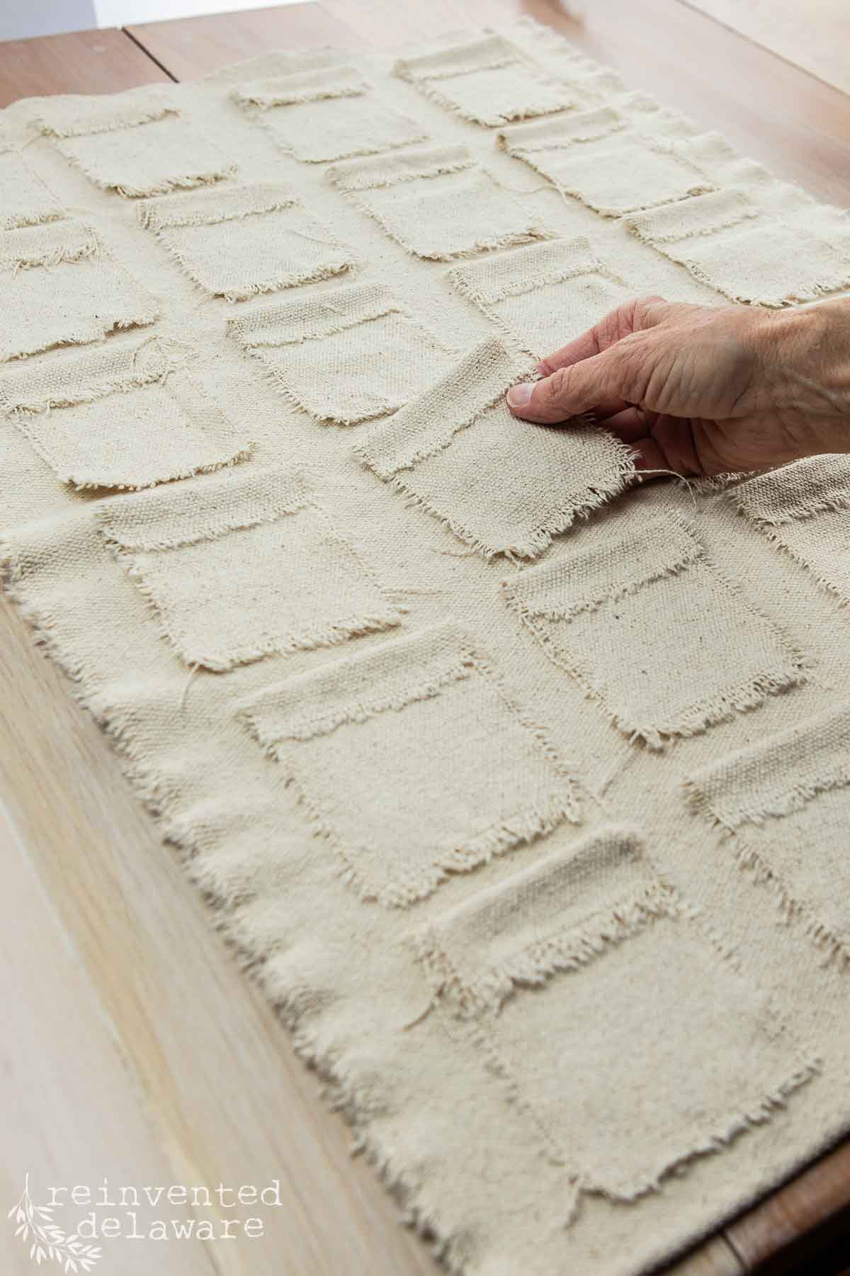 A hand lifts one of many small, frayed fabric squares sewn onto a larger rectangular fabric piece spread on a wooden table. The fabric squares are arranged in rows and columns. The text “reinvented delaware” is in the bottom left corner.